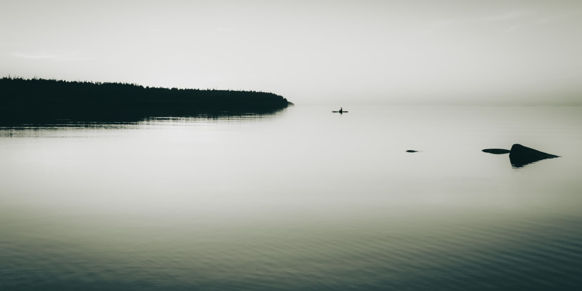 Stillt, monokromt sjölandskap med skog, sten och en ensam paddlare